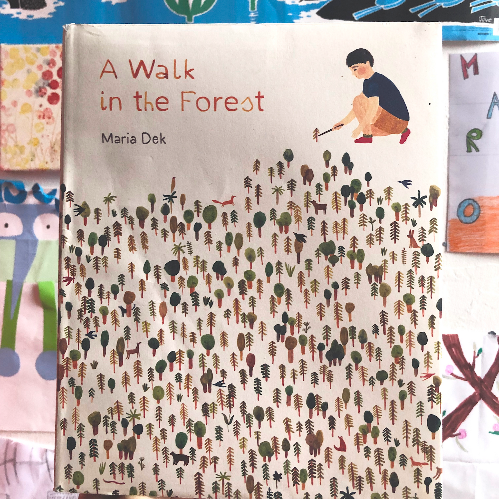 A book cover is in the foreground of the image. The title  A Walk in the Forest is painted  in brown on the cover A painted boy in blue t-shit and brown shorts kneals beside the title text. Underneath both is a forest of small painted trees. Behind the book cover is an out of focus wall coloured posters and children's drawings 